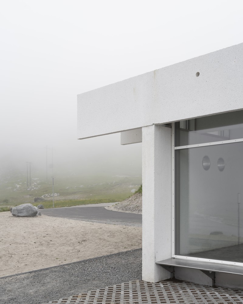 Foto av hjørne på hvitt servicebygg, vei, fjell og skyer i bakgrunnen.