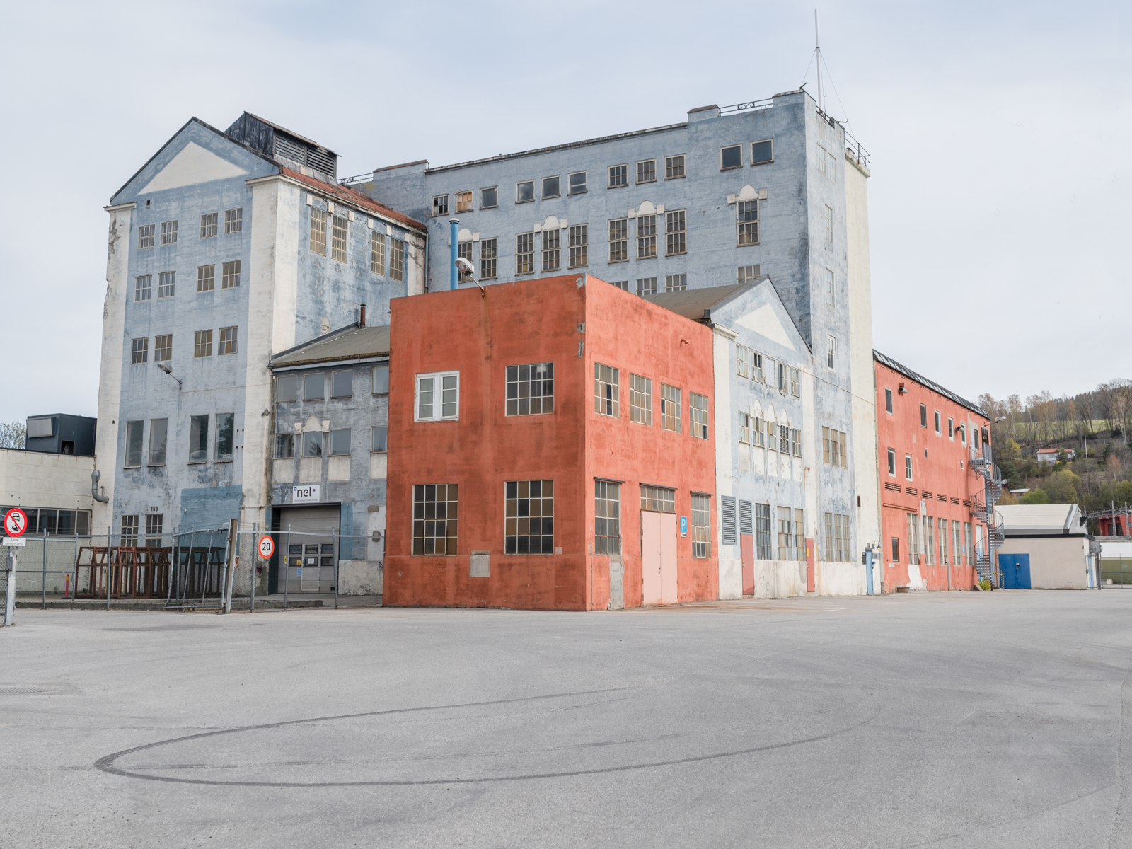 Foto av rødt og lyseblått industribygg, asfaltplass i forgrunnen.
