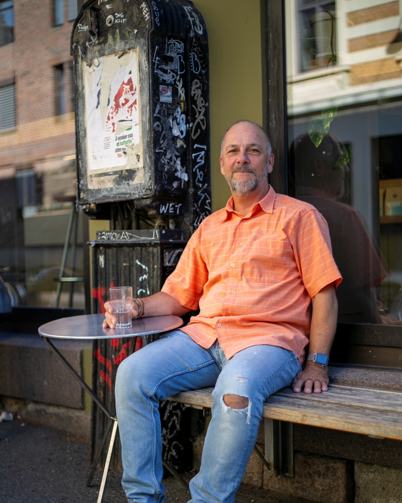 Foto av Jeremy Williams som sitter på en benk utenfor en kafe med et glass i hånda.