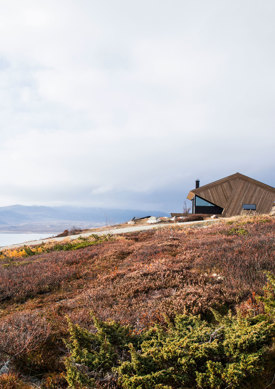 Hytte sett på avstand over lyng og med en fjord og fjell i bakgrunnen