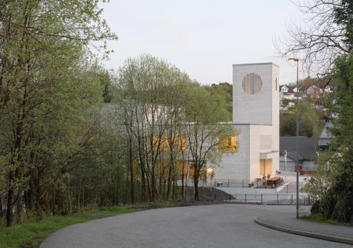 Foto av en vei som leder ned til en kirke i hvit teglstein, trær i forgrunnen.