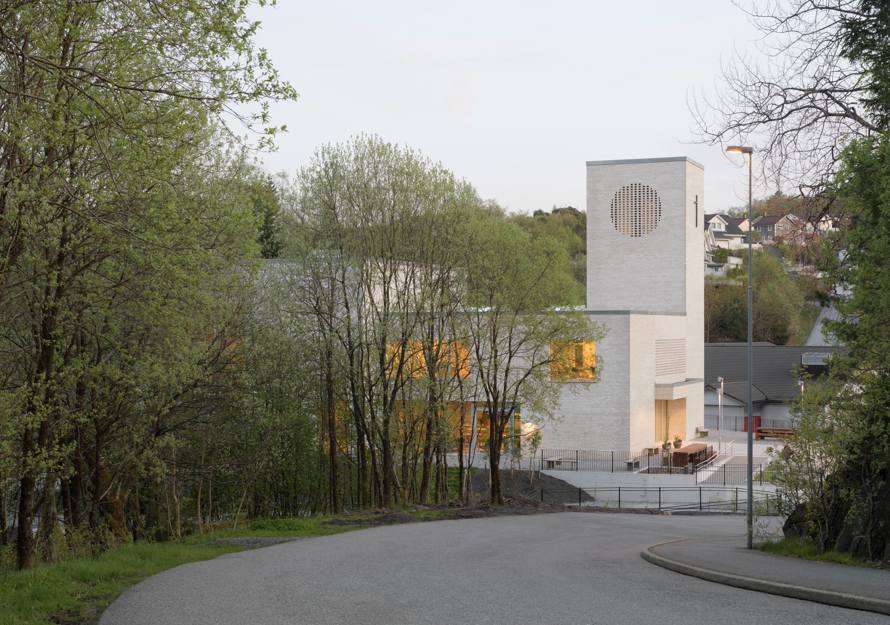 Foto av en vei som leder ned til en kirke i hvit teglstein, trær i forgrunnen.