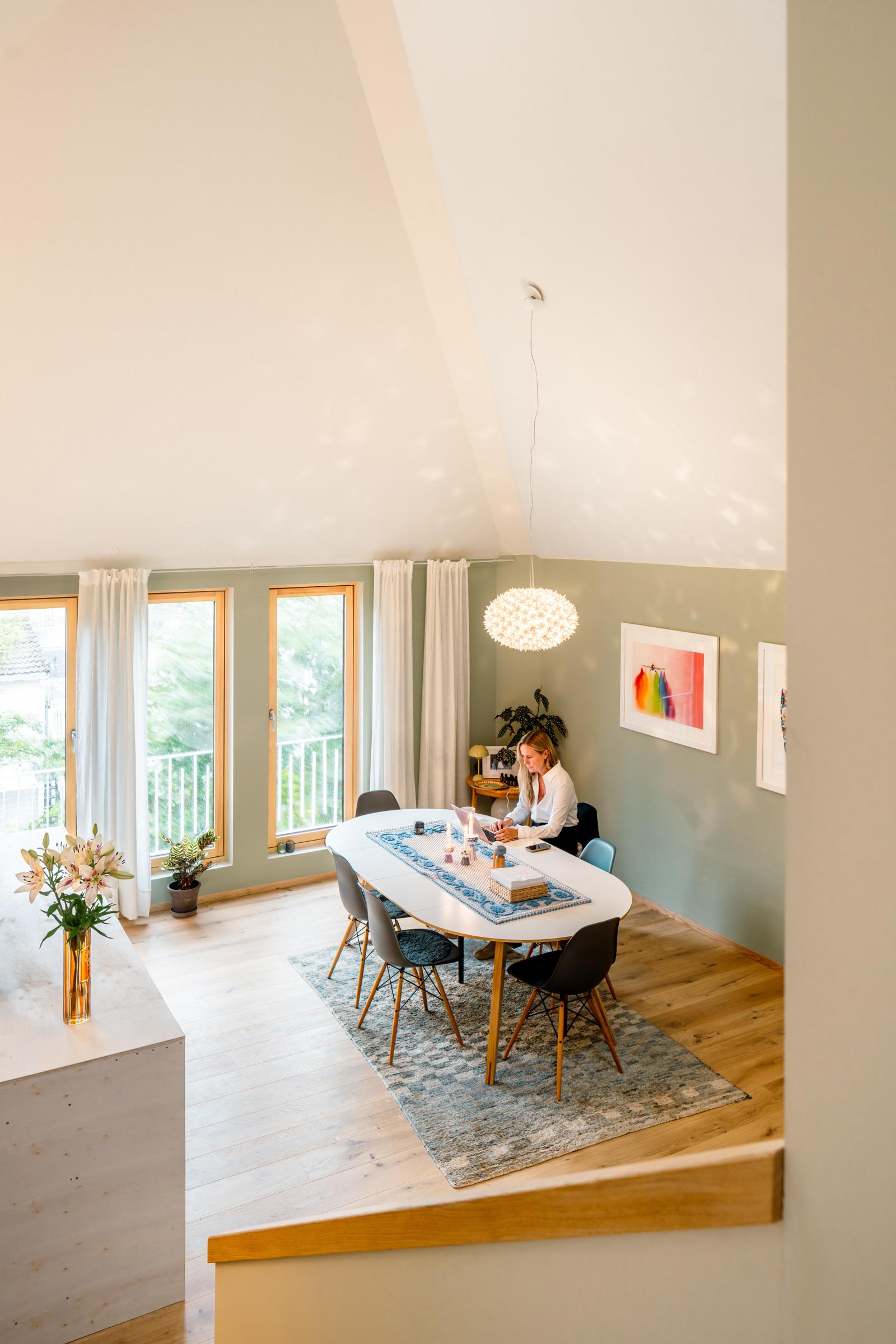 Interiør foto tatt fra trapp ned på stuebord hvor ei dame sitter på PC'en.