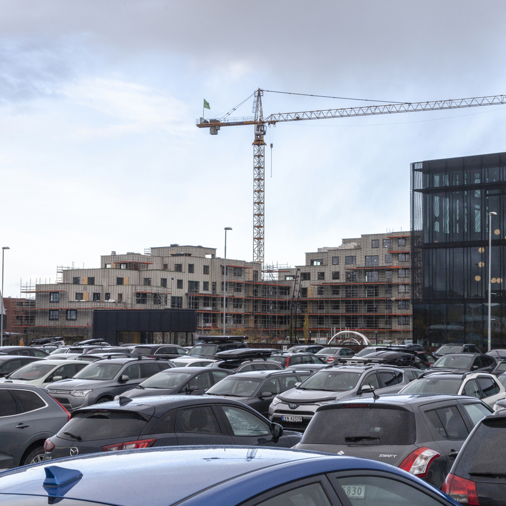 Parkeringsplass med mange biler, heisekran og bygg i bakgrunnen.