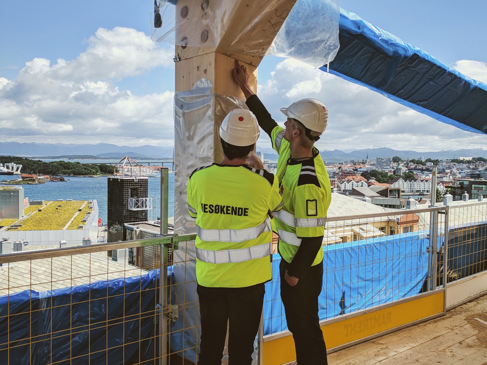 To arbeidere i gult ser på en forbindelse mellom to trelementer, en søyle og en bjelke. Foto.