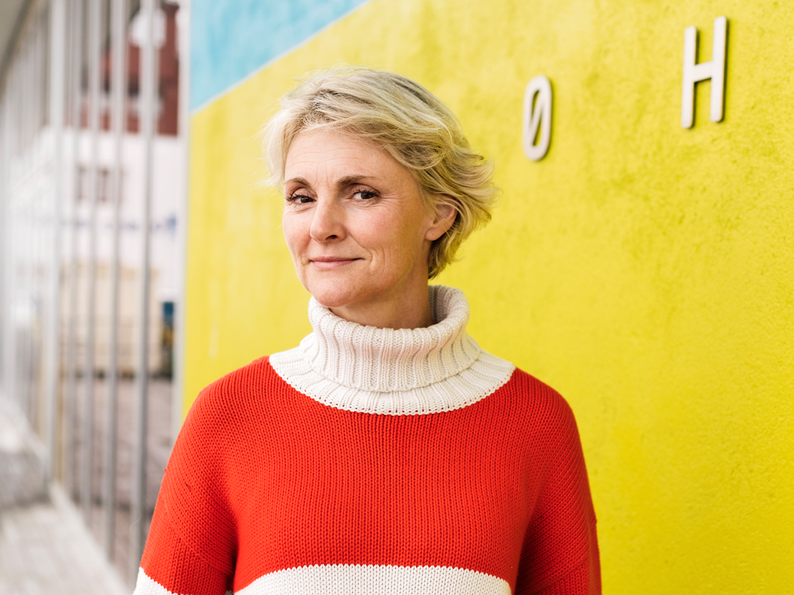 Foto av Tine Hegli i rød genser foran en knall gul vegg.