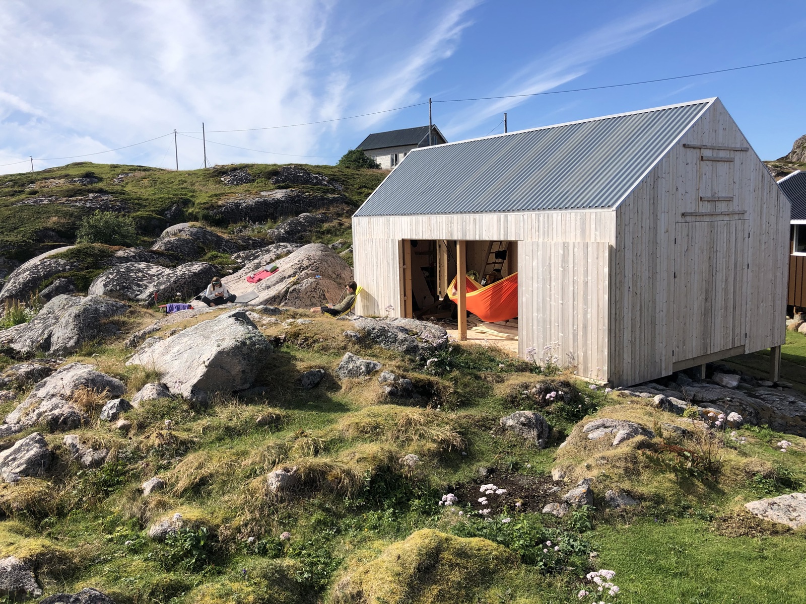 Foto av et naust i grånet treverk blant svaberg, hengekøye hengende i åpning.