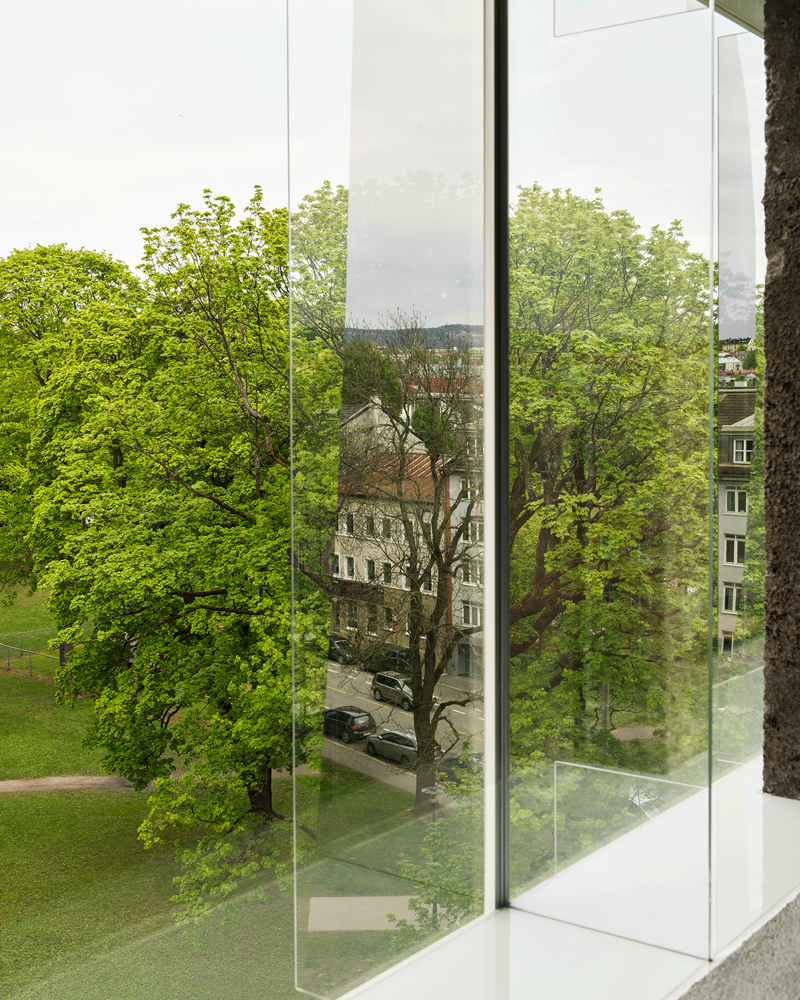 Fotografi av glassfasade med utstikkende glasselementer tatt fra innsiden ut til en park.