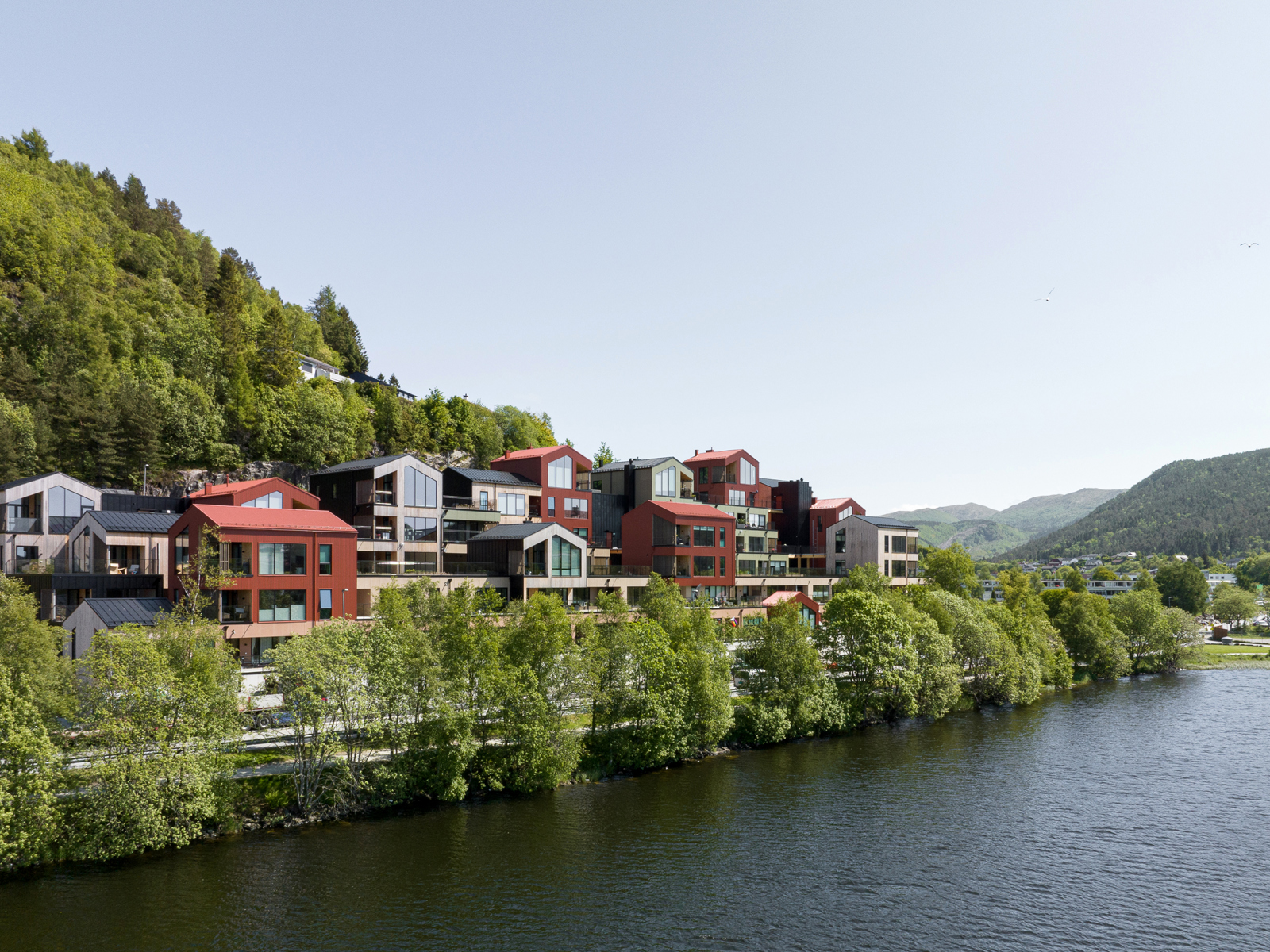 Oversiktbilde av boligkompleks i ulike farger i det grønne, vann i forgrunnen. Foto.