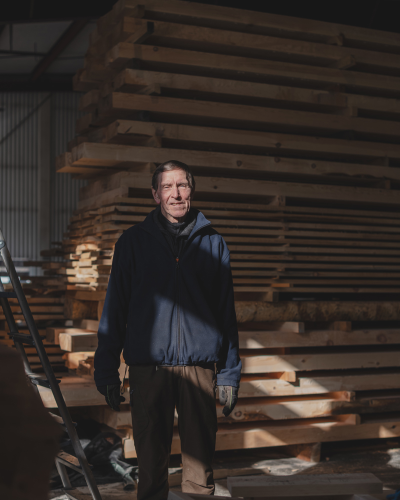 Foto av en mann som står i et lagerrom med masse treverk stablet bak seg.
