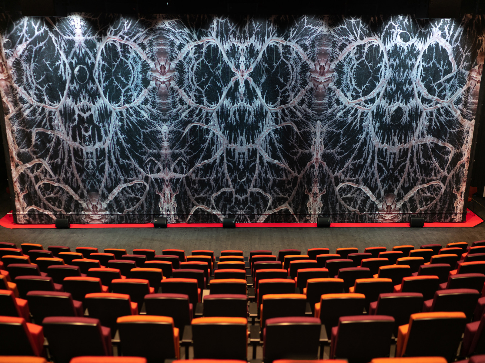 Foto av kunstneristisk sceneteppe, auditorium stoler i forgrunnen.
