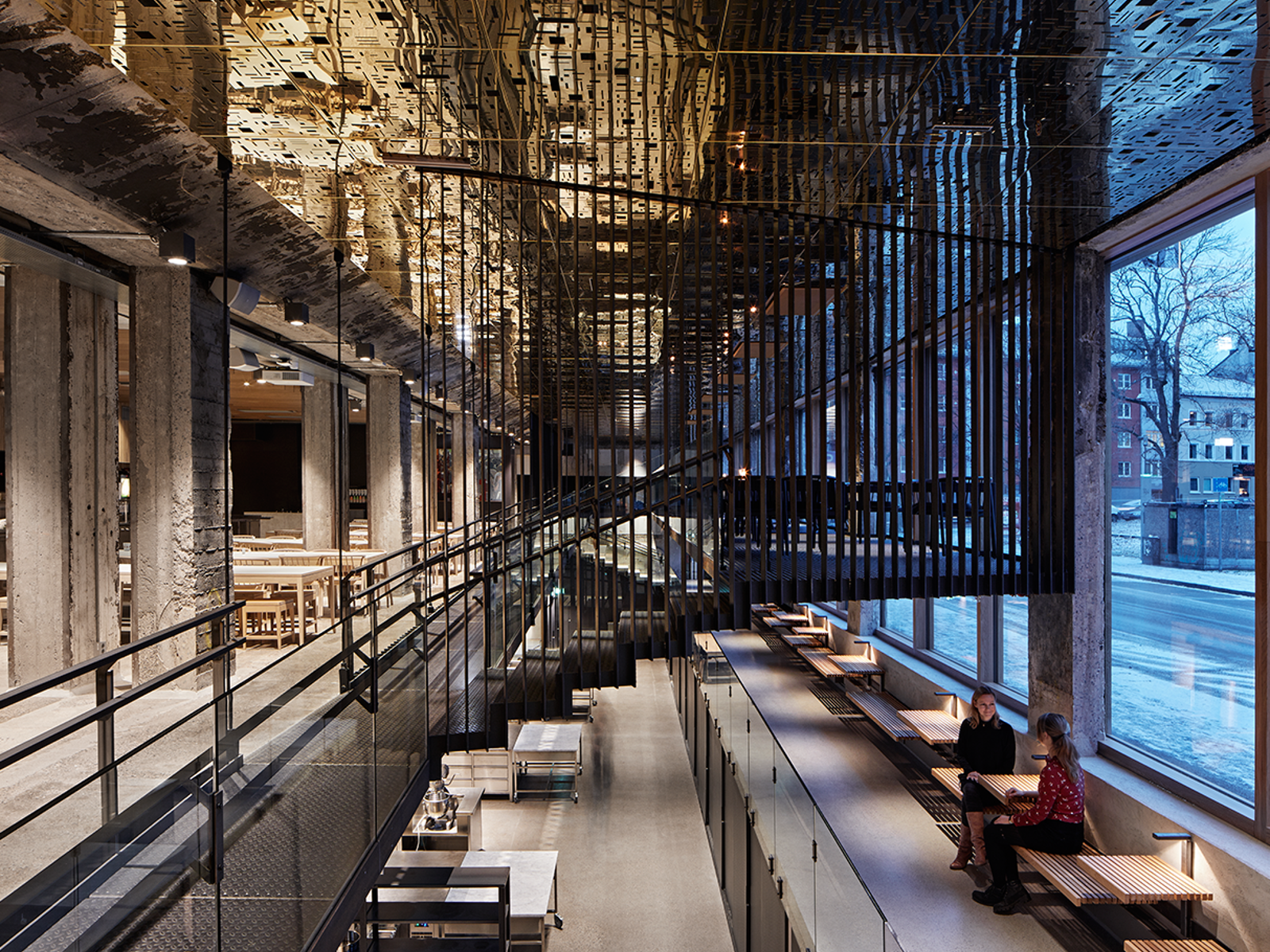 Interiør fotografi av bygg med flere sittegrupper, åpent ned til etasjen under, reflekterende gullplater i taket, ståltrapp med spiler i midten.