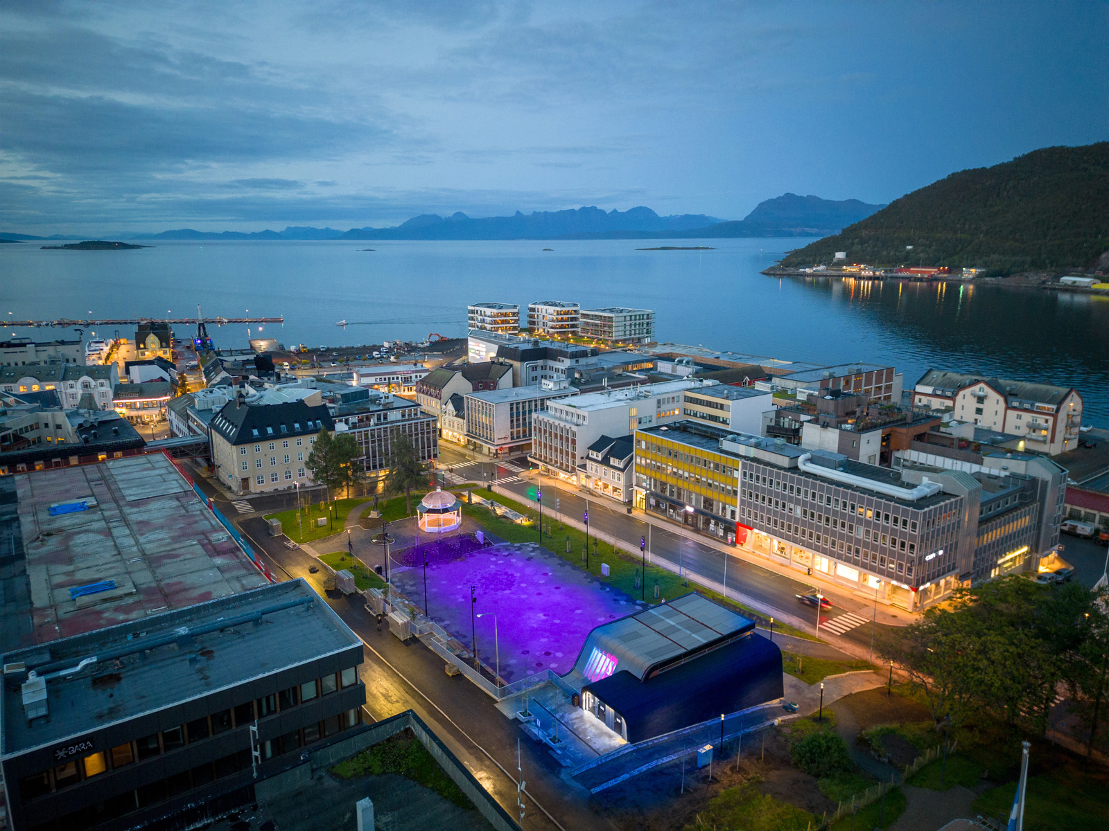 Oversiktsfot, dronefoto, av utendørs scene og opplyst plass i bykontekst, hav og fjell i bakgrunnen. 
