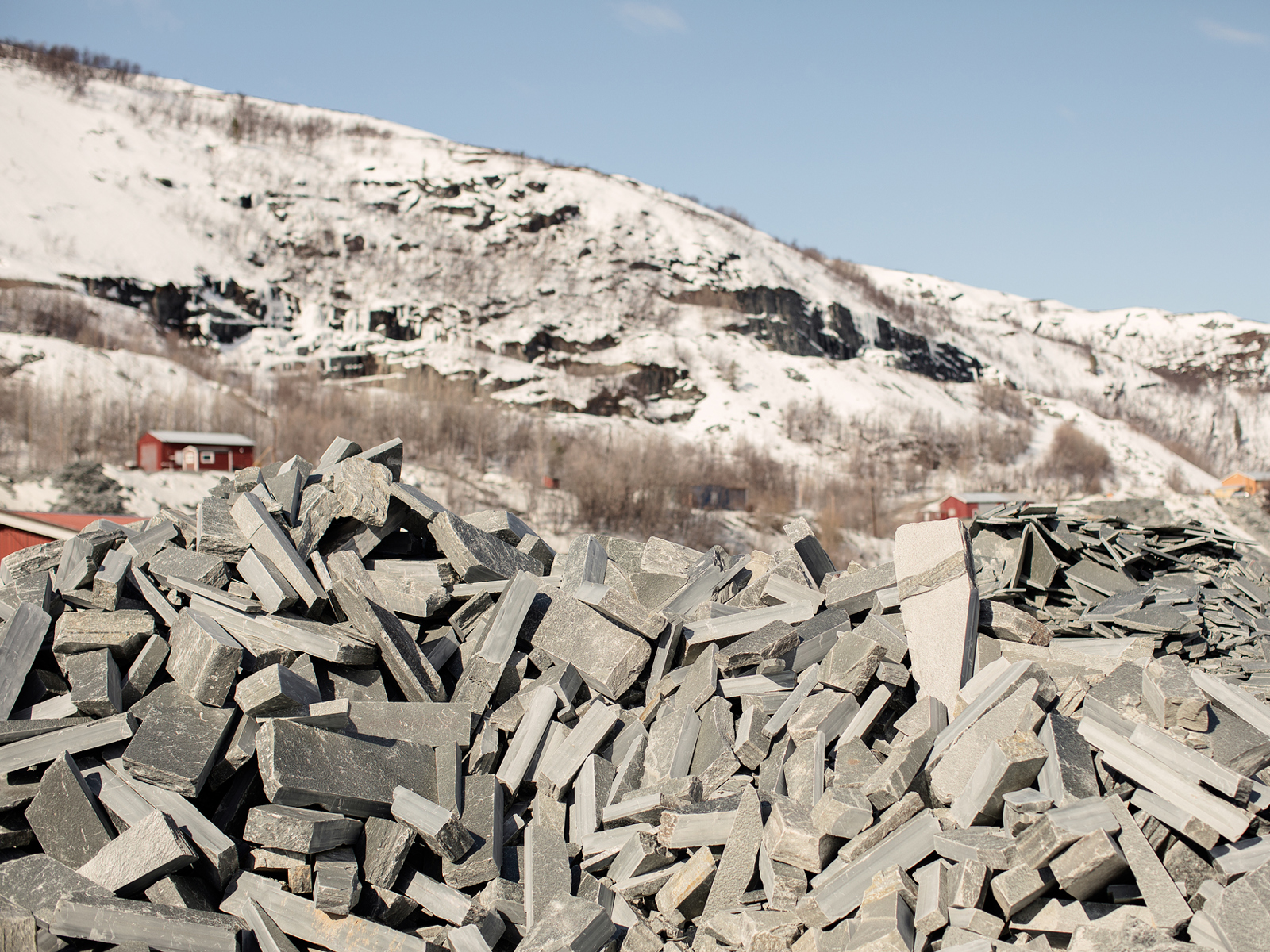 Foto av hauger med skifer utendørs, snøkledd fjell i bakgrunnen.