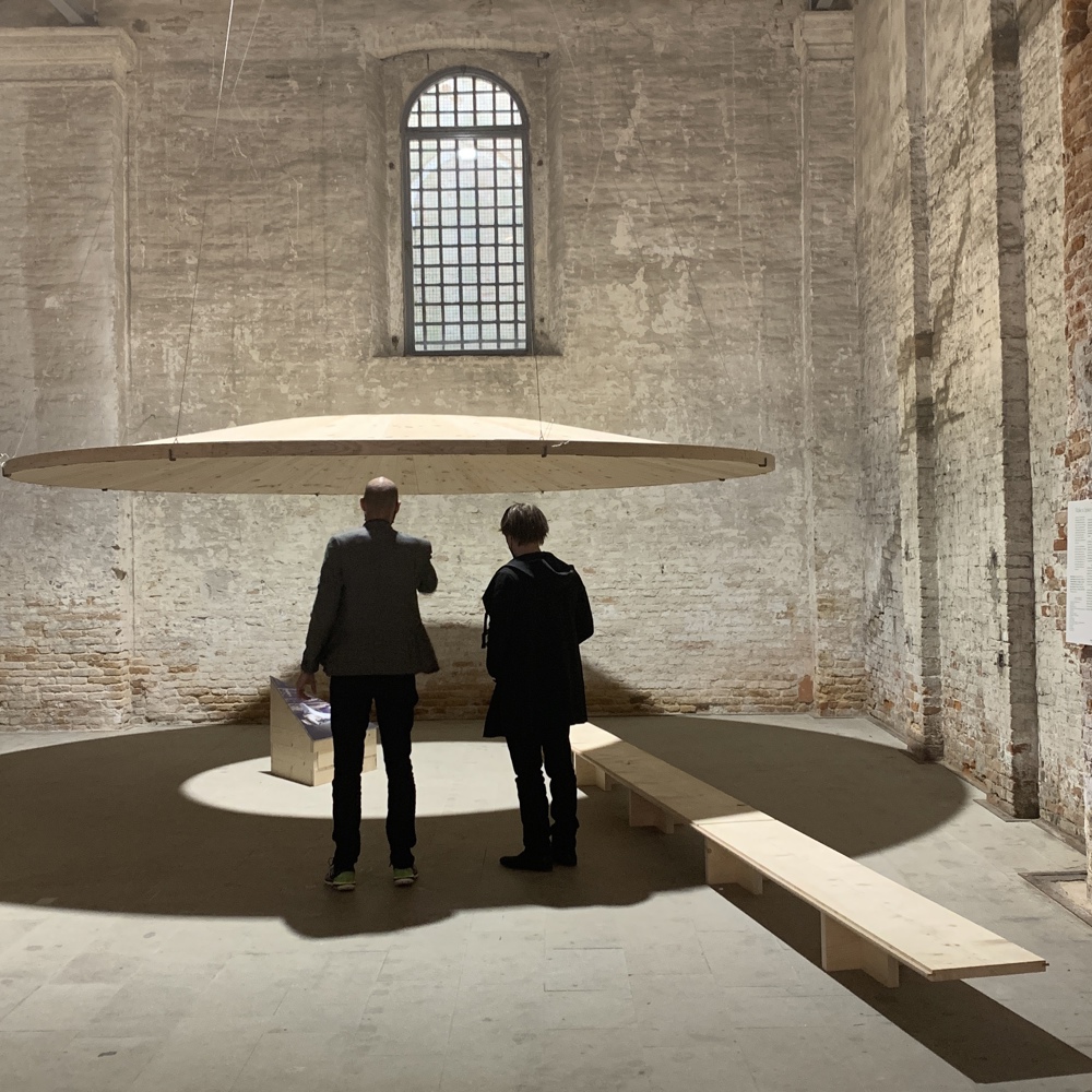 to mennesker står å ser på en utstilling av en rund tredisk hengende i lufta med hull i midten. Foto.
