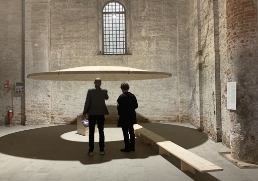 to mennesker står å ser på en utstilling av en rund tredisk hengende i lufta med hull i midten. Foto.