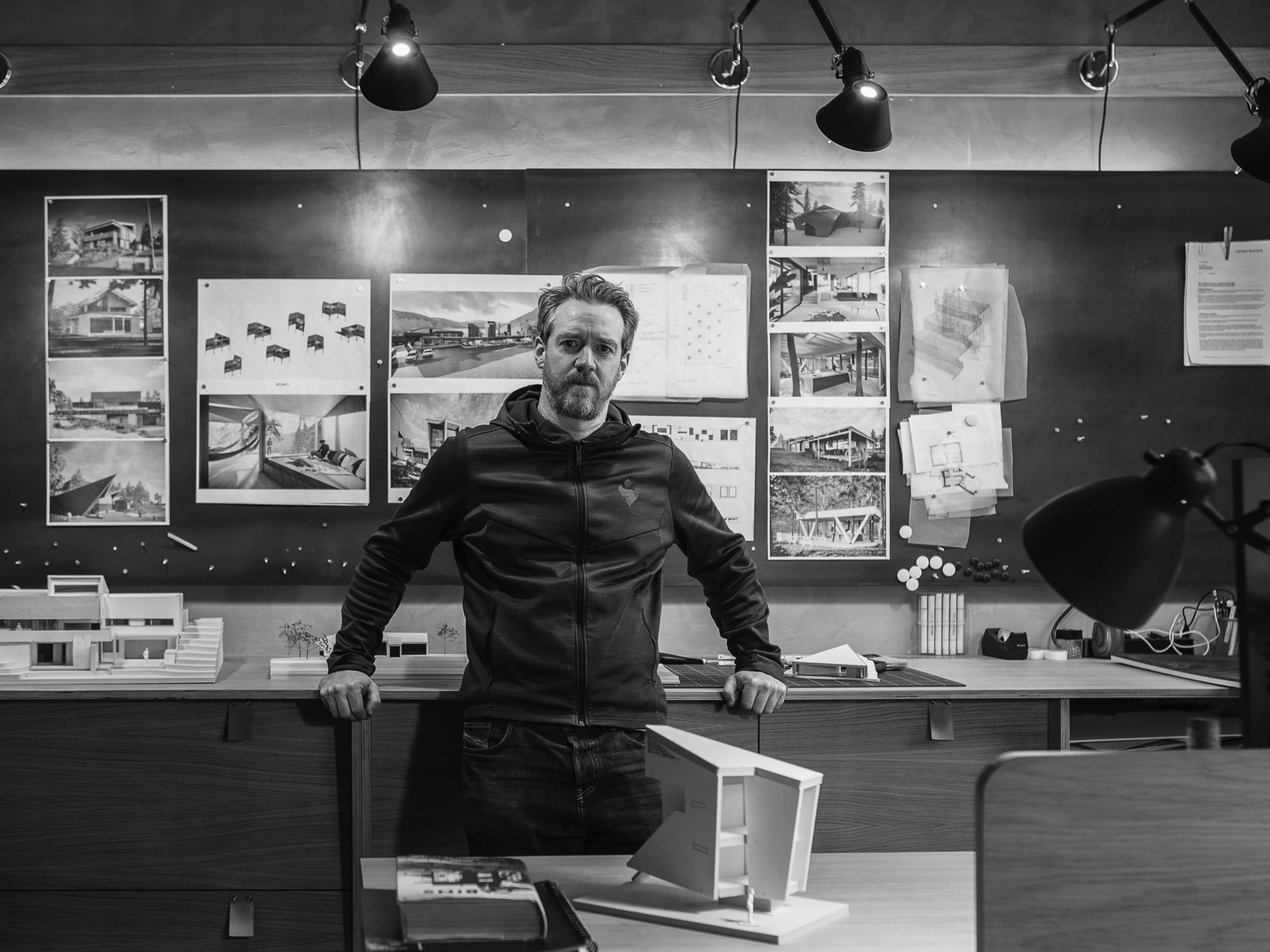 Mann står og lener seg inntil skrivebord på et arkitektkontor. Svart hvitt foto.