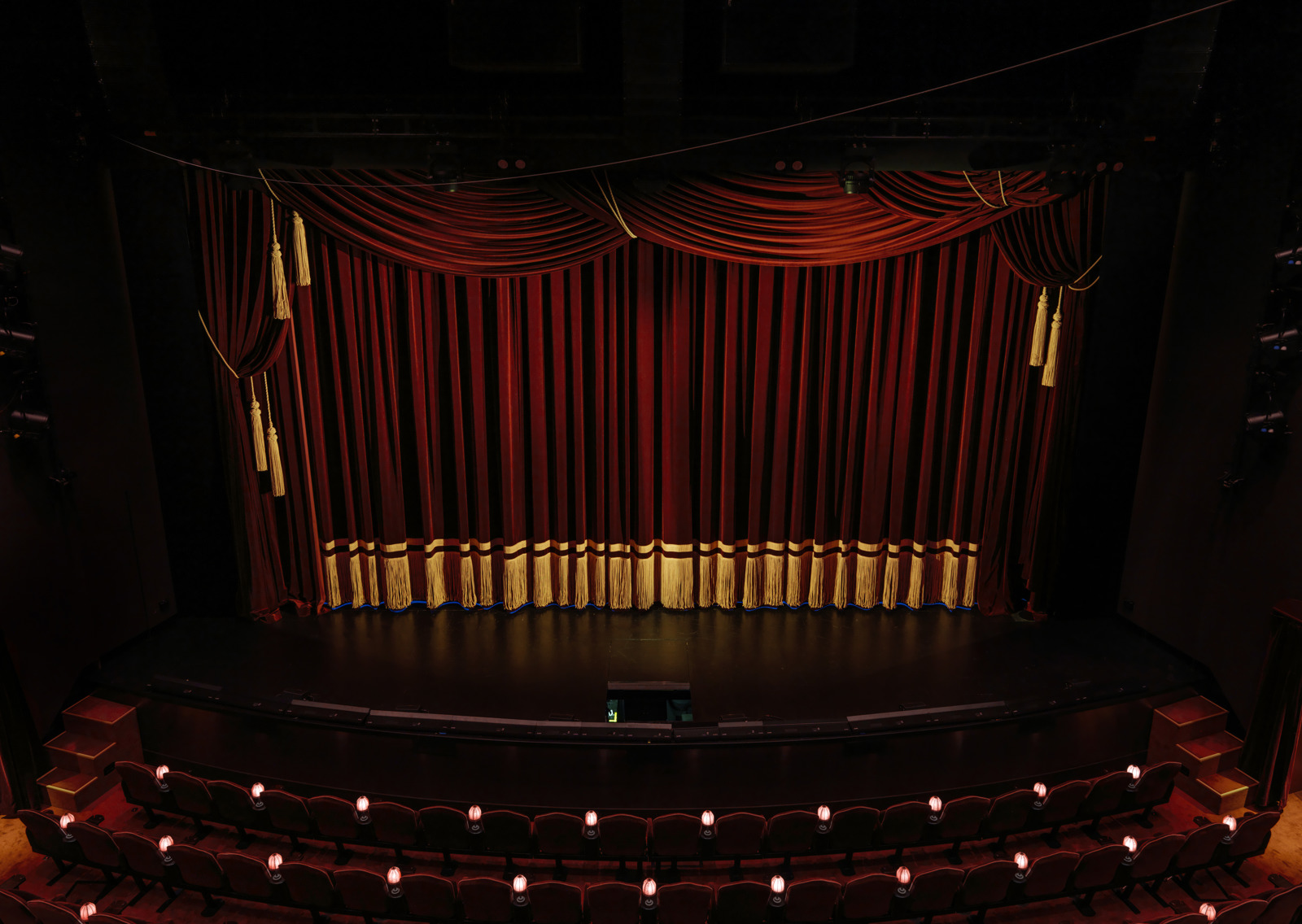 Interiørfoto av teatersalen i nye hjorten teater, røde scenetepper er trekt for og små lamper lyser i salen.