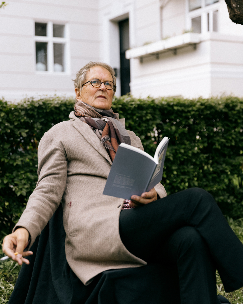 Thomas Thiis Evensen sitter på en stol ute, leser en bok og røyker. Foto.