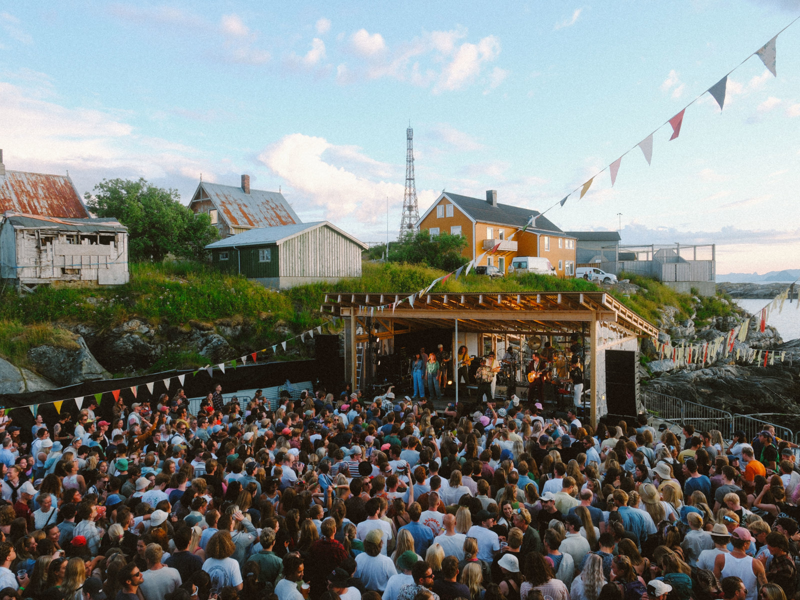 Foto av konsert utendørs i sommerkveldinga. Masse mennesker, vimpler, enebolig i bakgrunnen.