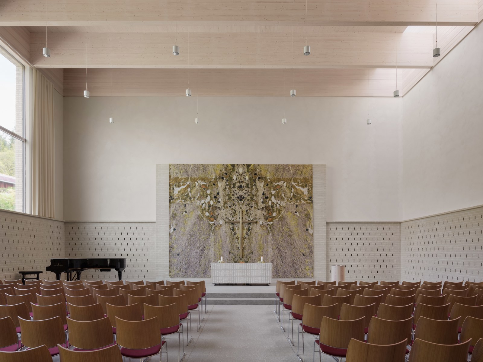 Foto av et lyst kirkerom sett mot alteret, rader med stoler, piano og et stort kunstverk bak alteret.