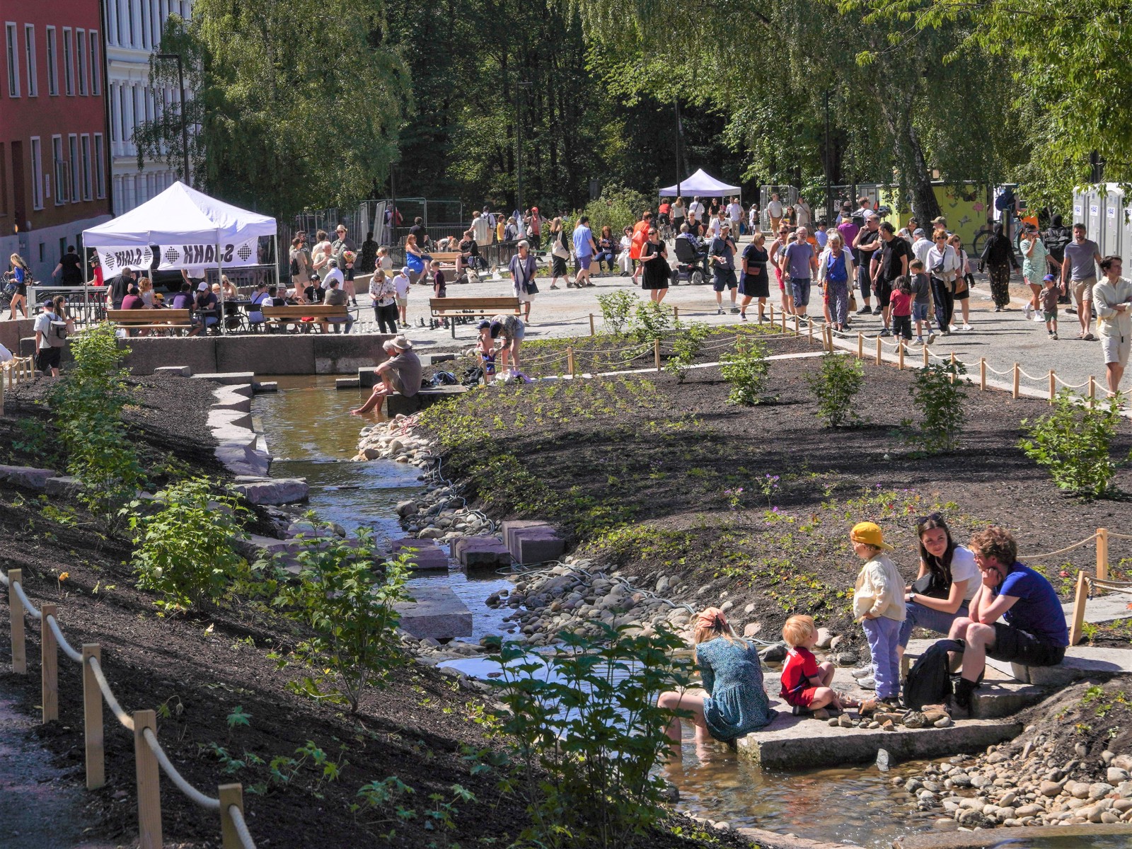 Mennesker oppholder seg langs en bekk, på gangvei, i bykontekst. Foto.
