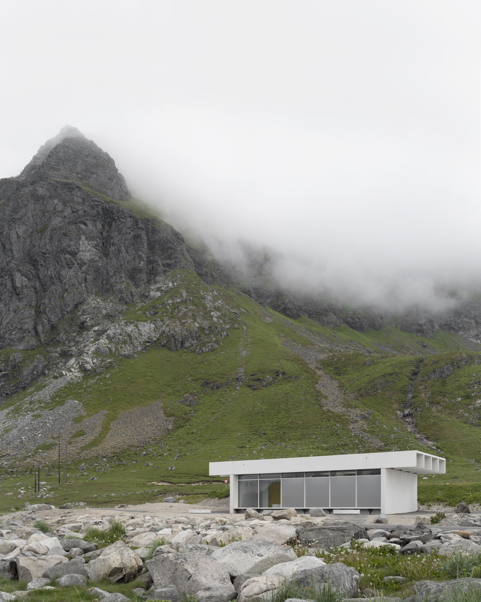 Foto av lite hvitt servicebygg midt i landskapet, fjell og skyer i bakgrunnen. 