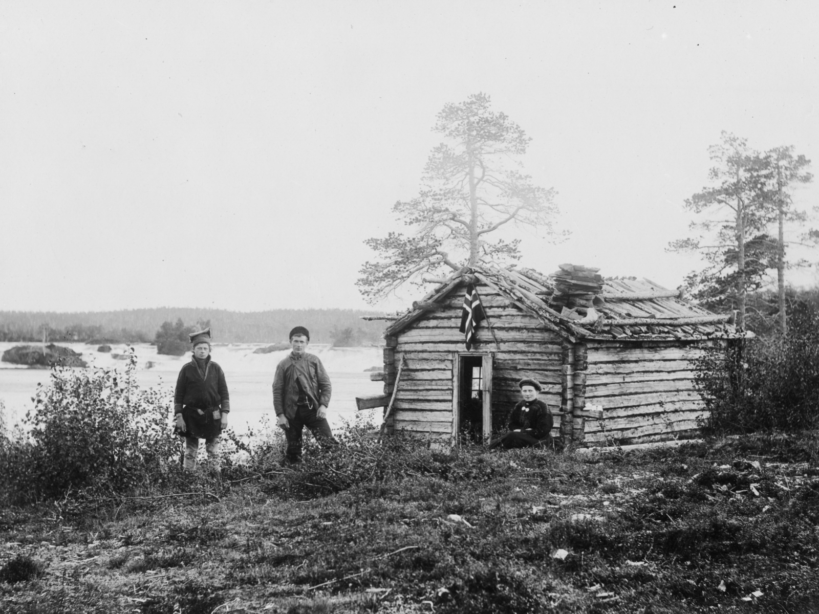 Historisk bilde i svart hvitt av tømret bygg med tre mennesker utenfor. Foto.