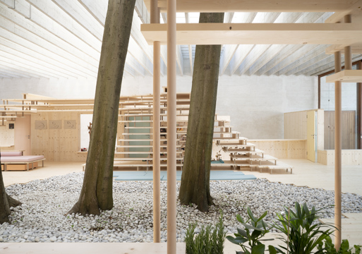 Den norske paviljongen i Venezia, tre trær som går opp igjennom taket, taket består av mange bjelker, gulvet av grus der treerne står og hardt dekke ellers, betong vegger. Trekonstruksjoner preger interiøret. Foto.