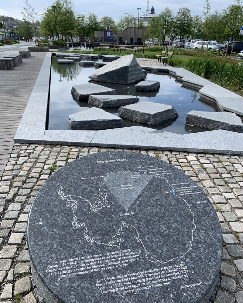 Parkområde, firkantet vanndam med steiner i, stein med informasjon i front.