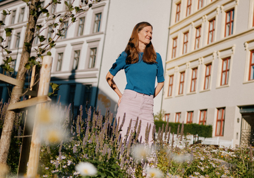 Foto av Sally O'Halloran stående, i forgrunnen er det blomster og planter, i bakgrunnen bygårder.