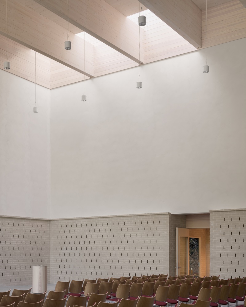 Interiør foto i kirke, rader med stoler, lys vegg i tegl og puss, limtrebjelker i taket og overlys fra takvinduer.