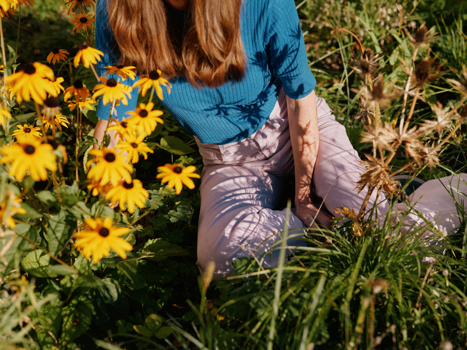 Foto av en kvinne med rødt hår, blå genser og rosa bukse sittende blant blomster.