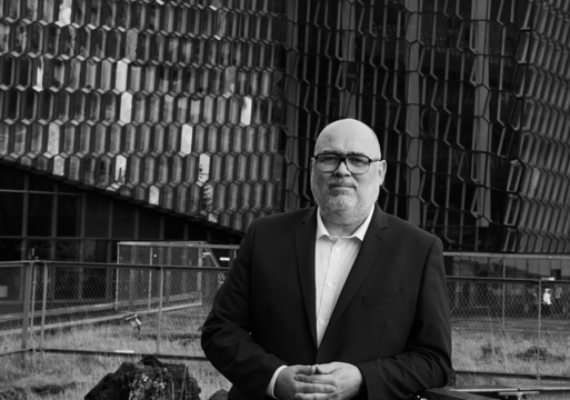 Portrett foto av Logi Már Einarsson med et stort bygg i bakgrunnen, svart hvitt.