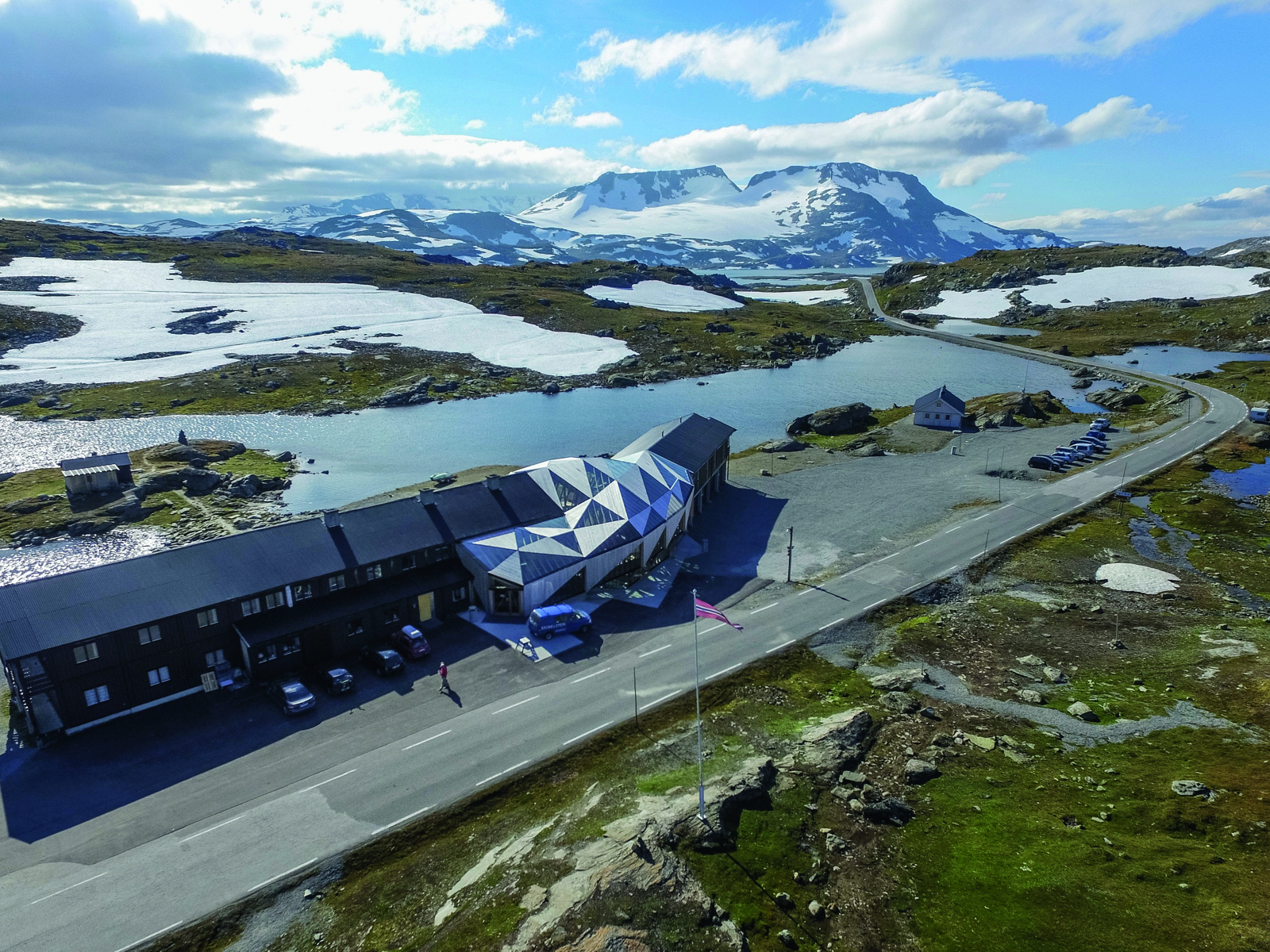 Overblikksbilde av Sognefjellshytta i landskap med vei, vann og fjell med snø på i bakgrunnen. Foto.