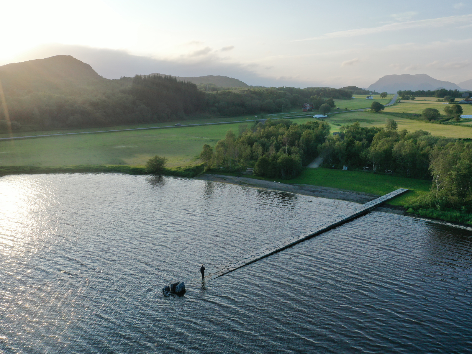 Foto ovenfra som viser en steinlagt sti eller forre som går ut i vannet og sakte forsvinner under vannflaten, en mann står på enden. skog, fjell og grønne jorder rundt vannet.