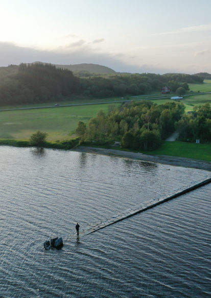 Foto ovenfra som viser en steinlagt sti eller forre som går ut i vannet og sakte forsvinner under vannflaten, en mann står på enden. skog, fjell og grønne jorder rundt vannet.
