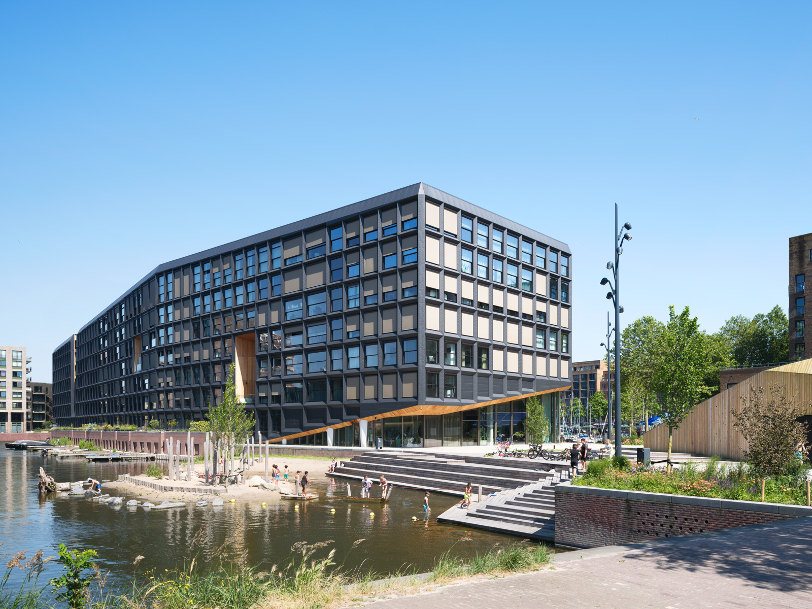 Foto av fasade på boligprosjekt, vann og uteområde i forgrunnen, barn som leker. 