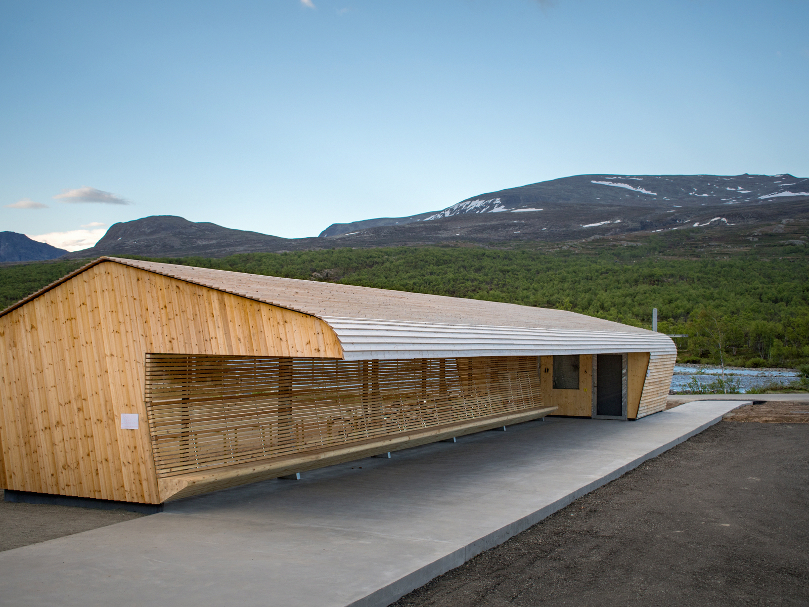 Reinsvangen servicebygg, Overbygg i treverk med integrerte sittebenker, runde former på tak. Fjell i bakgrunn. Foto.