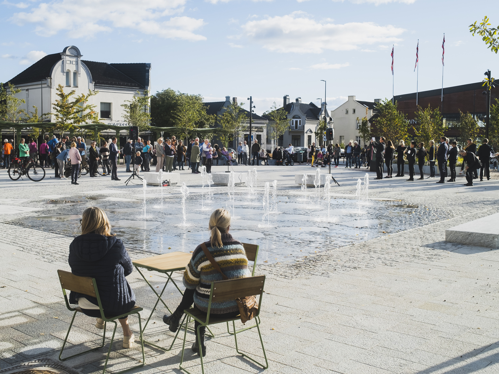 Torg med mange mennesker, fontener og vann på midten, to damer sitter på noen stoler ved et bord og ser ut på torget. Foto.