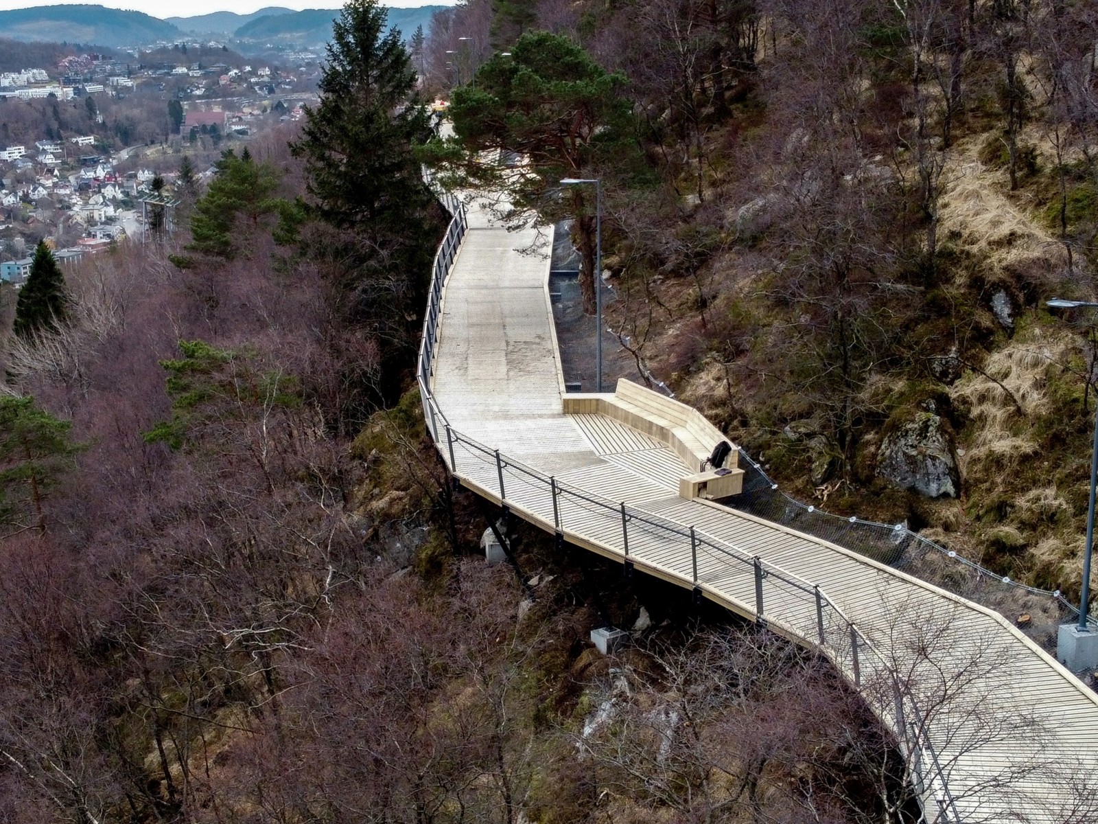 Oversikts foto/ dronefoto av gangsti i treverk i åsside, med by i bakgrunnen.