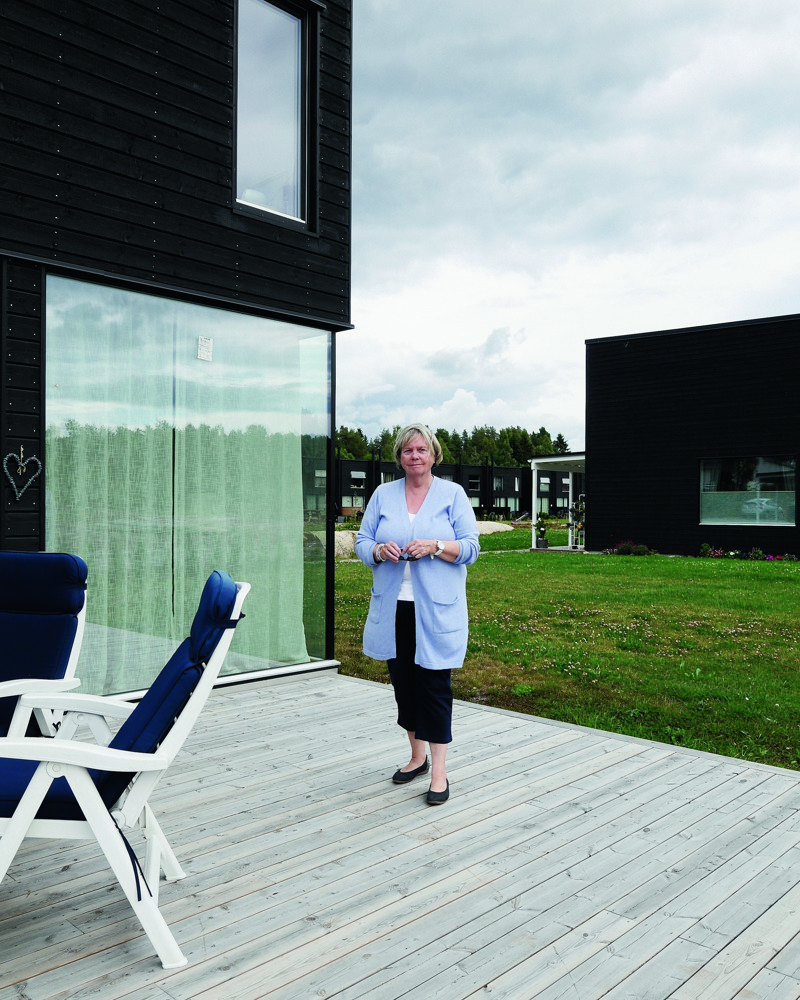 En eldre dame står på en treplatting utenfor et rekkehus, flere rekkehus og skog i bakgrunnen. Foto.