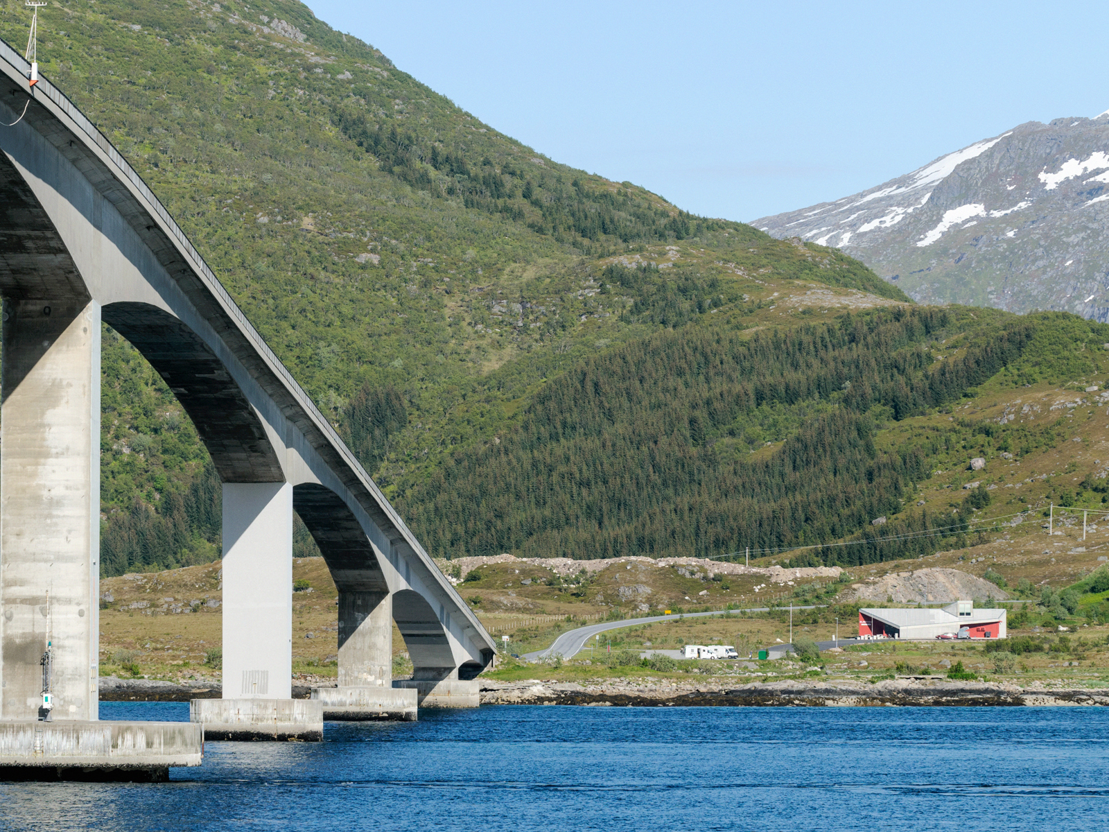 Betong bro som fører over vann til grønn natur og vei, slake buer under broa. Foto.