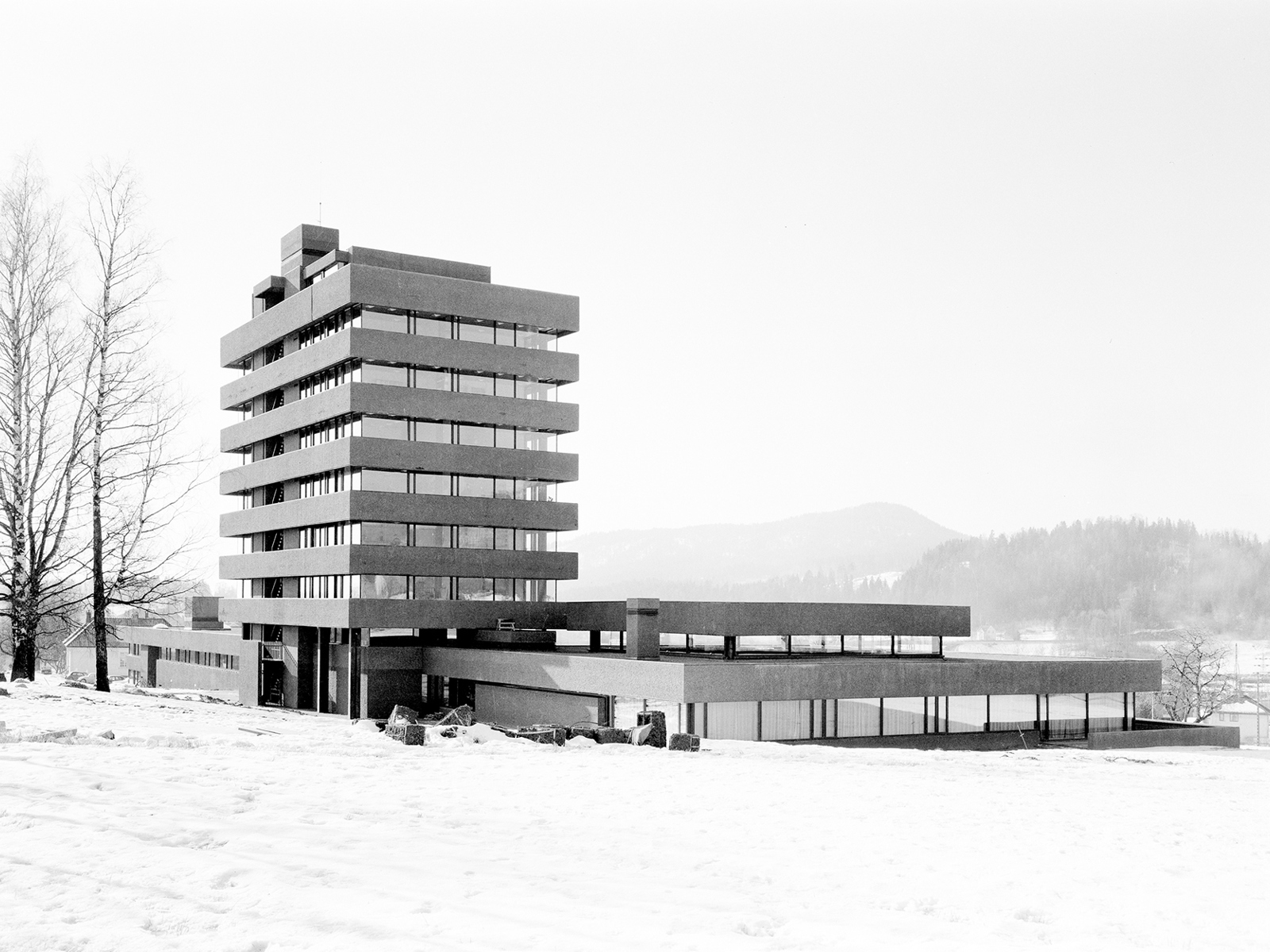 Modernistisk bygg på 7 etasjer står i et snølandskap, svart hvitt. Foto.