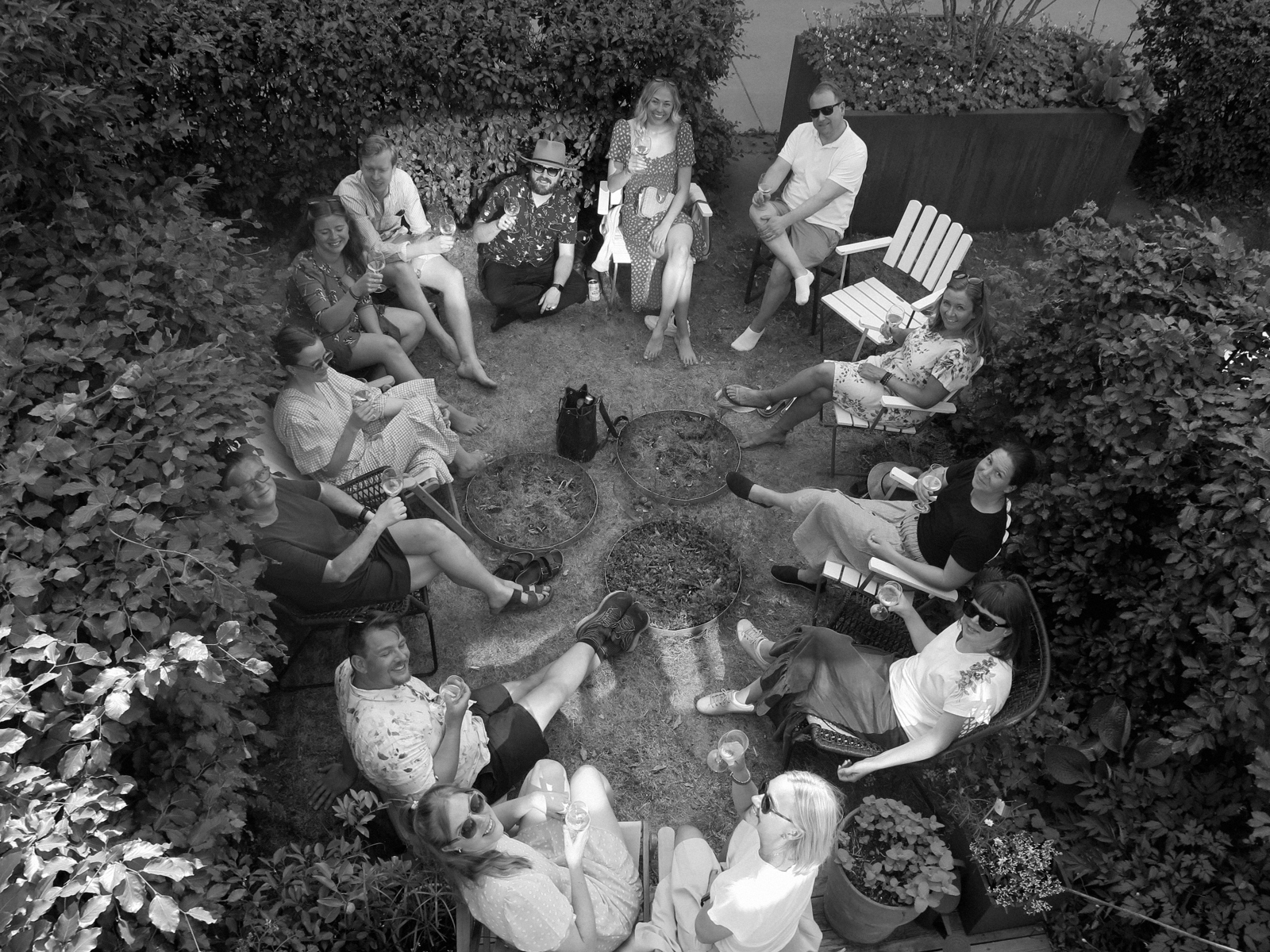 Mennesker sitter i en sirkel i en hage med busker rundt og ser opp på fotograf. Foto.