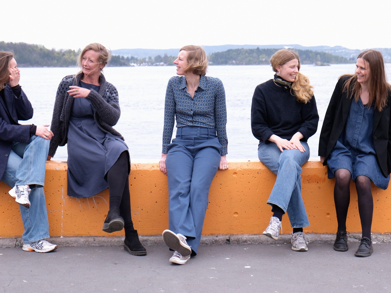 Fem mennesker sitter og snakker på en oransje bryggekant, vann i bakgrunnen. Foto.