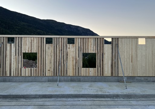 Foto av fjøs under bygging, en vegg av ombruks tre, fjell i bakgrunnen. 