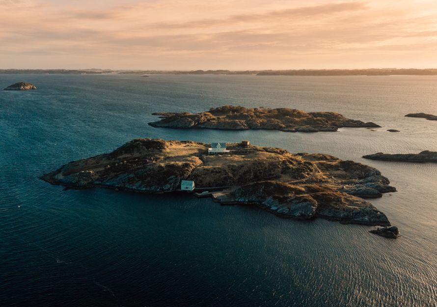Drone foto av en øy med to hus på, hav og mindre øyer rundt.