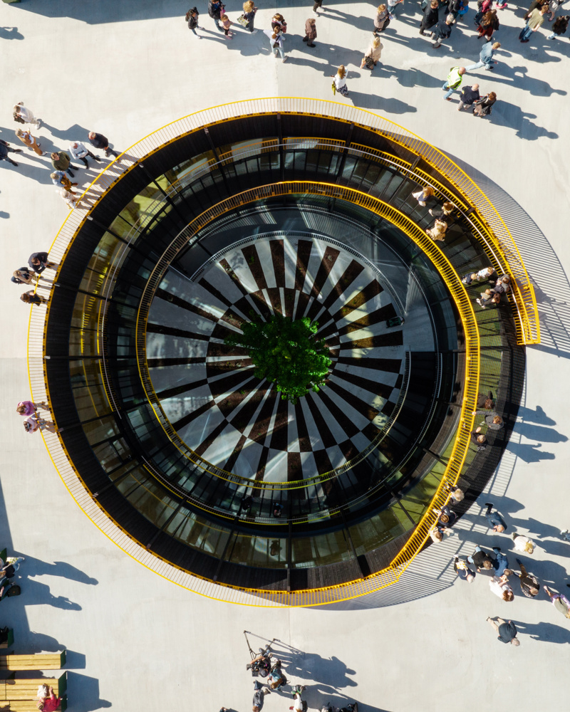 Fly/dronefotografi av tak med et sirkulært hull med trapp ned i midten. Mennesker går på taket. Foto.