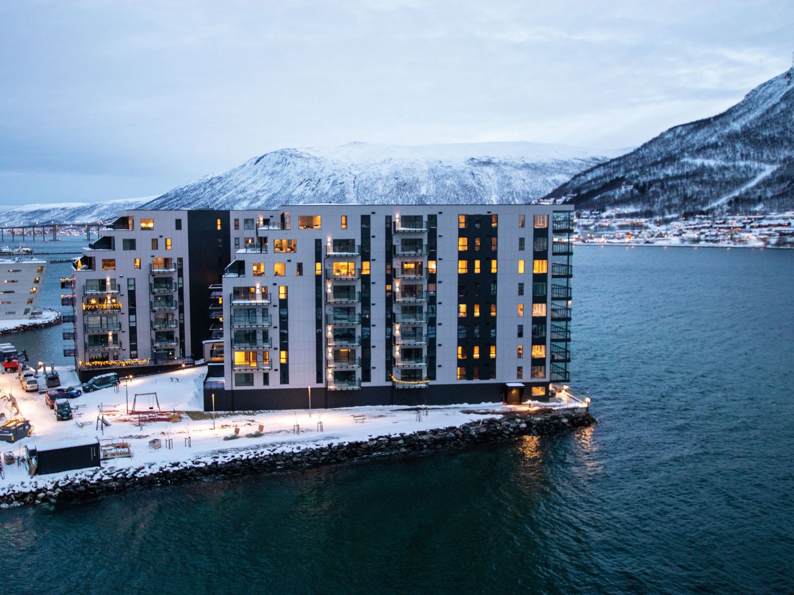 Foto av boligblokk på tuppen av havn, vann rundt, snøkledde fjell i bakgrunnen, vinter lys. 